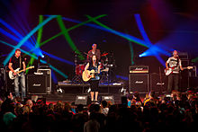 A photograph of four people standing on a stage in front of a crowd of people looking up at them with green and blue swirls in the background