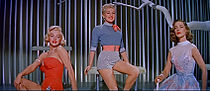 Monroe in How to Marry a Millionaire. She is wearing an orange swimsuit and is seated next to Betty Grable, who is wearing shorts and a shirt, and Lauren Bacall, who is wearing a blue dress.