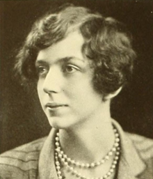 A young white woman with wavy hair cut in a side-parted bob, wearing three strands of beads and a collared jacket in a plaid or striped fabric