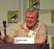 A man with white hair, glasses, and a brown shirt sits at a table with a microphone; a card in front of him reads "Mike Carlin".