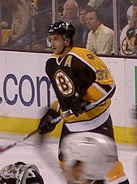 Hockey player in black and yellow uniform with a large "B" in the middle. His mouth is slightly open and his stick is raised diagonally off the ground.