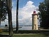 A small pale yellow metal lighthouse with a red roof on the shore of a lake with a massive white cloud behind