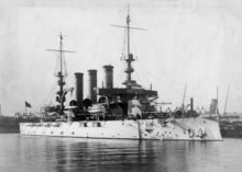 A white battleship with three smokestacks and two tall masts sitting in port.