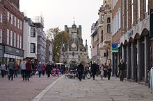 A view down a pedestrianised street with walkers and shops each side with a sixteenth century buttercross in the distance