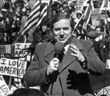 Photo of Jerry Falwell holding a microphone at a political rally. American flags and people holding "I Love America" signs are visible behind him.