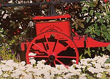 Photographie d'une charrette rouge et fleurie datant 1930 selon l'inscription écrite en noir, entourée de géraniums et de plantes d'exposition.