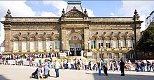 A queue of people at the edge of a pedestrianised square stretches uphill across the scene from right to left, then doubles back up another slope to the double-arched entrance at the centre of a large mid-nineteenth-century stone building. This also has steps leading up to the entrance with four ornamental street-lamps in front. On either side of the entrance are giant pilasters, two more of which can be seen, surmounted by urns, at the building's corners. There are six large windows with shell tympanums and scroll-effect balcony rails between the entrance and the corners on each side, with five small circular windows above and between the tympanums. Below on each side are basement windows, those on the left obscured because of the sloping site. Above the entrance is a large arch and above that, in gold lettering, is "Leeds City Museum". Higher up is another gold inscription, "Leeds Institute". Above this is another, larger, tympanum with sculpture, set in a pediment with urns on either side. Behind is a mansard roof.