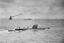 A submarine sailing on the surface of the ocean, while in the background is a large warship and a smaller vessel.