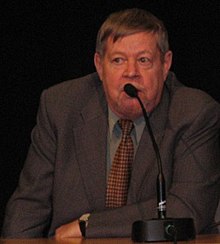 Color photo: Medium shot of Arto Paasilinna, sitting behind a table, speaking in a microphone.