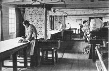 Black and white photograph of a factory room. Two men are hand-printing fabric on long tables. The fabric hangs from the ceiling at the end of the tables, displaying the patterns.