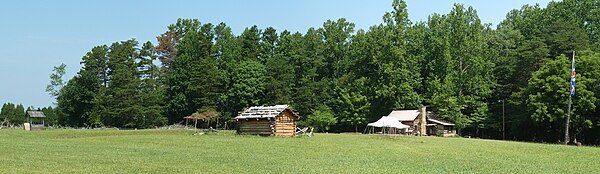 A panoramic view of the historic site showing two cabins and a well, as well as a flagpole bearing the King's Colours