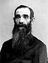 A man with a long dark beard and a dark shirt sits and looks straight ahead