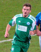 Short-haired white man wearing green and white sports kit, running on grass.