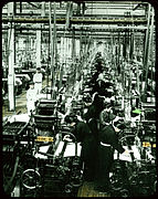 Photo de l'intérieur d'une usine. Alignées à perte de vue, des machines sont en fonctionnement, contrôlées par des ouvrières.