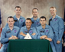 Photographie en couleur de six hommes en combinaison de vol posant avec une maquette de capsule Apollo.