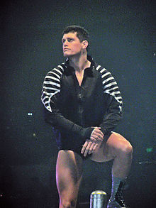 A caucasian male with black hair stands on the turnbuckles of a wrestling ring, with on foot on the top rope and the other on the middle rope, while looking to his right. He is wearing short black wrestling tights and black and silver wrestling boots, with a black jacket featuring a silver design on both sleeves.