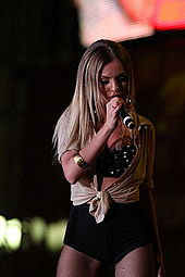 A blonde-haired woman is seen wearing a beige-blackoutfit as she performs to an audience.