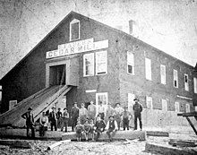 A black and white photograph of a large building featuring a sign that reads "E Faber's Cedar Mill"; More than a dozen white men sit on a large cedar log in the foreground