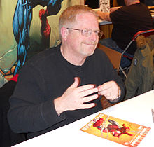 Man with black shirt and glasses, sitting at a table at a comic-book convention