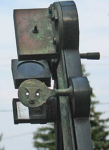 Octant details showing the double-holed sighting pinnula. Also visible is the small cover that can block one or the other of the holes. The horizon mirror is on the opposite side of the instrument. The left side is transparent while the tin amalgam on the mirrored side has completely corroded and no longer reflects light. The back of the index mirror's holder is at the top and the three circular glass shades in square frames are between the two mirrors. Octant pinnula and horizon mirror.jpg