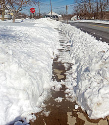 A sidewalk, covered with thin snow in many areas, with deep snow on the sides gouged irregularly. A stop sign is visible in the rear