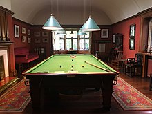 Photo of billiard room at Olveston Historic Home, with billiard table at centre, fireplace to left, overtable lights, skylight and various antiques
