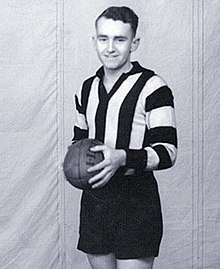 A dark-haired footballer in a long-sleeve black-and-white vertically-striped guernsey and black shorts, holding a football