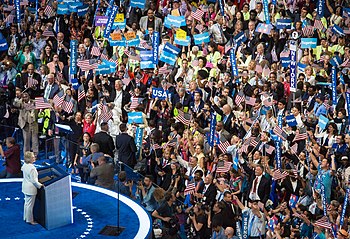 Вид сверху на синюю сцену. Женщина в белом брючном костюме стоит за трибуной (с прозрачными щитками) перед толпой сторонников с американскими флагами, знаками «Хиллари» и т. Д.