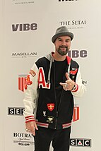 a man in a black trousers and a baseball jacket with a gray hat, standing on the red carpet