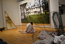Kramer is covered by a dirtied white sheet sitting on the floor. To her right is half of a canoe, and her left a rolled-up snow fence. Behind her is a large image of Pelican Lake residential school.