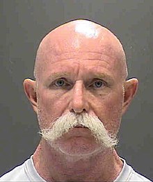 A mugshot of a bald white man with a white prospector's mustache; wearing a white t-shirt, he is facing and looking into the camera.