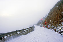 A snow-covered road next to a rocky cliff face on the right with only fog visible on the left. Ahead is a temporary traffic light showing red.