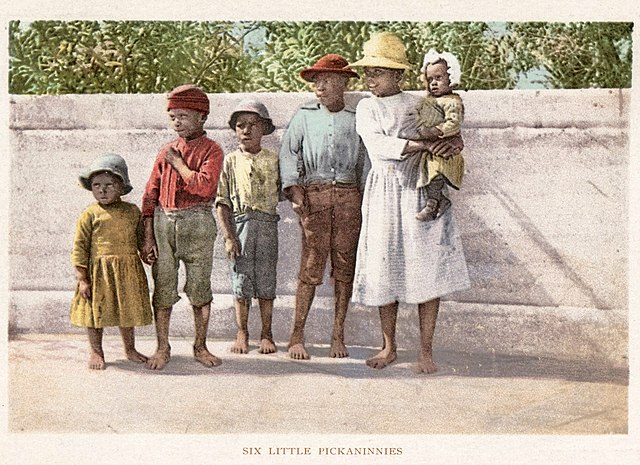 Postcard depicting a group of African-American children of varying ages, standing barefoot in front of a low wall with foliage visible behind it, all looking away from the camera to the left; the oldest child holds an infant on her hip