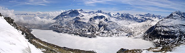 Il Lago del Moncenisio ghiacciato, contornato dalle montagne, visto dalle vicinanze del Col de la Tomba (parte del Gruppo Roncia-Lamet). Sulla sinistra la Val di Susa, coperta dalle nuvole, dalle quali spuntano vette del Gruppo dell'Orsiera. Sullo sfondo sono riconoscibili (da sinistra verso destra) la Cima di Bard (3.168 m), il Monte Giusalet (3.313 m), il Monte Malamot (o Pointe Droset) 2.917 m, i Denti d'Ambin (3.372 m), la Punta Sommeiller (3.333 m), la Rognosa d'Etiache (3.382 m) (fra le due montagne il Colle del Sommeiller a 2.993 m s.l.m.), Le Petit Vallon (3.236 m), la Roche d‘Etache (3083m) (con davanti il Colle del Piccolo Moncenisio, la Pierre Menue (o Aiguille de Scolette) (3.506 m), e la Punta Clairy o (Signal du Petit Mont Cenis) (3.162 m). Tutte le montagne sono parte del Gruppo d'Ambin, tranne la Pierre Menue che appartiene all'omonimo gruppo.