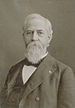 Head and shoulders of an older white man with a bushy beard wearing a suit coat, vest, and bow tie.