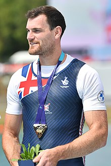 Aldridge trägt einen dunkelblauen Rennanzug mit einem Union Jack an der rechten Schulter über einem weißen T-Shirt. Die Goldmedaille ist an einem blauen Band um seinen Hals befestigt. In den Händen hält er einen Blumentopf.