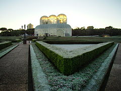 A hedge fence's corner is covered in light frost. In the distance, the Botanical Garden's greenhouse can be seen.