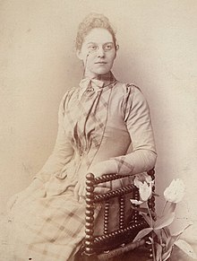 Black and white photograph of a woman, Florence Taylor the astronomer, seated in a chair, wearing a checked close fitting high-necked dress and pince-nez. There is a vase of tulips next to her.