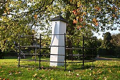 White painted village pump within wrought iron fence, under an autumnal maple, with shed leaves on the surrounding grass.