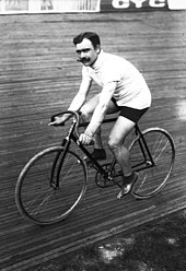 A black-and-white photograph of a man with three-colored sweater and shorts with a moustache sitting on a bicycle.