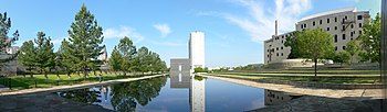 A panoramic view of the memorial. In the center is a large stone structure shaped as a gate with "9:03" at the top. At the center of the gate is a large hole and through it a road can be seen. The Regency Towers building is visible on the right of the image in the background. The gate is reflecting in a pool of water in front of it, and grass and trees are visible to the left and right of the pool.