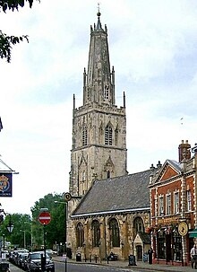 A stone church in a town seen from the southeast. St the far end is a large tower surmounted by a truncated spire with pinnacles and, at the top, a ball finial