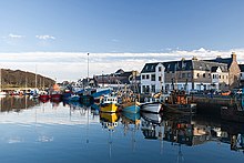 Still blue water in the foreground with numerous small coloured boats along a harbour and buildings against a blue sky in the background