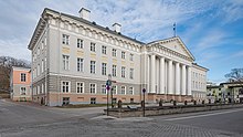 Seitliche Farbfotografie eines barocken, dreigeschossigen Gebäudes mit einem weißen Säuleneingang. Unter dem Dach verzieren einfache, weiße Ornamente die hellgraue Fassade. An der linken Wand neben dem Eingang ist „Tartu Ülikool“ eingemeißelt.