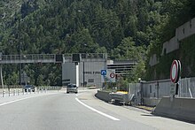 Photographie, depuis la route, de l'entrée d'un tunnel où plusieurs automobiles circulent, entouré d'une forêt dense, avec des panneaux routiers de circulation ; il y a le nom du tunnel d'Orelle inscrit en grandes lettres sur le bâtiment.