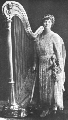 A white woman standing with a harp, wearing a long gown