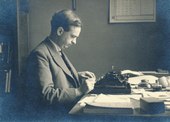 Middle-aged Hendrik Coenraad Blöte using a type writer. Hendrik is wearing a suit, tie and jacket. Many papers, some books and a jar of ink are on the desk.