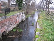 View downstream of the lock chamber build for the Sleaford Navigation, to maintain the head at the mill. The chamber is largely brick built with stone details for load-bearing parts, and the brick is coloured with moss and lichen. A little desultory grass covers the top sides. There are no lower gates, the lock having been converted into a weir many years ago. A cheap iron railing fence, painted black recently, delineates the property associated with the mill and restaurant to the left. This is a winter view and many bare trees line the banks downstream. The trunks of the nearest can be clearly seen to be covered in ivy. The water looks clear and placid.