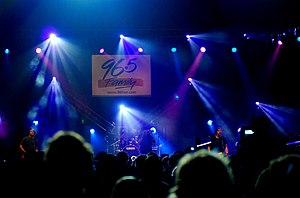 Project 86 performs at a concert. The entire band plays on a stage in front of a group of people, engulfed in blue lights.