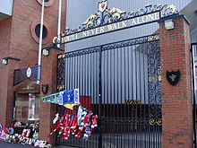 A set of gates with the inscription "You'll Never Walk Alone" above them. Flowers are wrapped into the gate and by the side of the gate